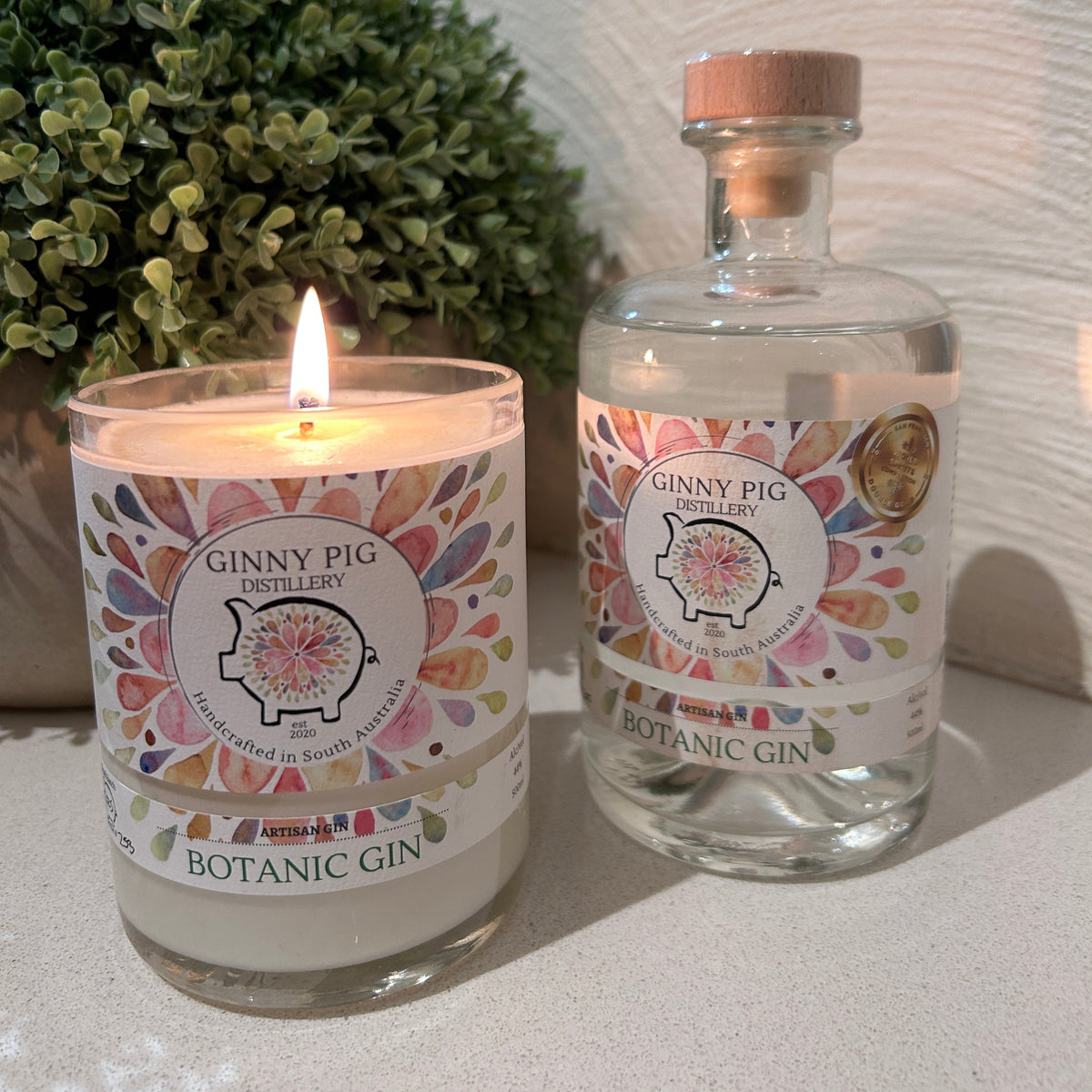 A lit glass jar candle and a clear glass bottle of Ginny Pig Botanic Gin with a cork stopper, both featuring colorful floral labels, set on a light surface with a green plant and textured wall in the background.