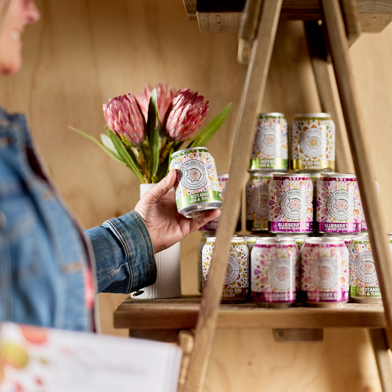 Person holding a can of gin next to a wooden stand with more cans and flowers.